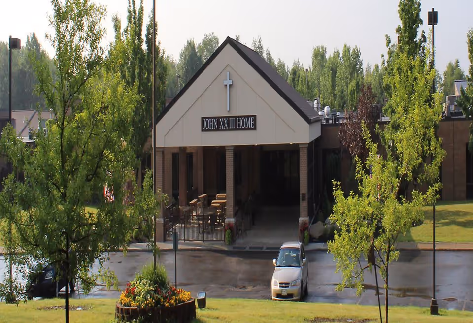 Front exterior view of the Saint John XXIII Home building with a peaked roof entrance, a cross above the entrance, surrounded by trees and a parking area with a few cars.