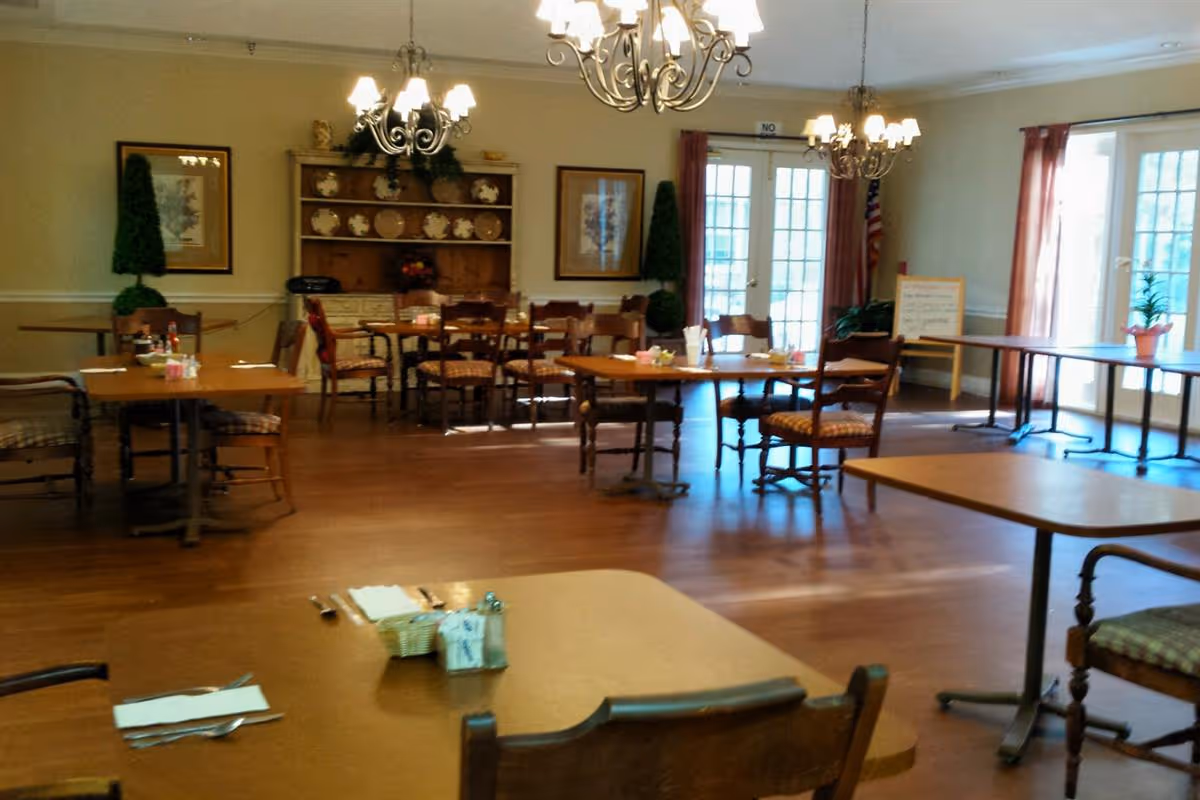 A dining room in Columbus Oaks Healthcare Community with multiple wooden tables and chairs arranged neatly. The room features hardwood flooring, chandeliers hanging from the ceiling, framed artwork on the walls, and a wooden hutch displaying decorative plates. Large windows and glass doors with curtains allow natural light to enter the space.