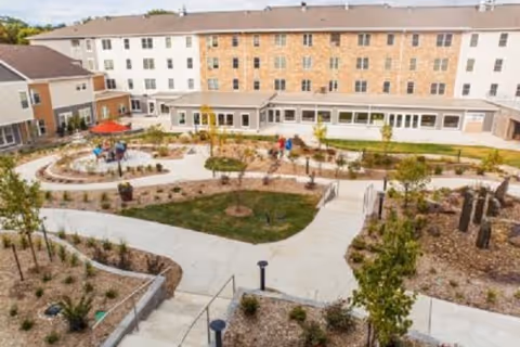 View of an outdoor garden area with walking paths, small trees, and landscaped plants in front of a multi-story senior living facility building. There are a few people walking and sitting under a red umbrella at a table.