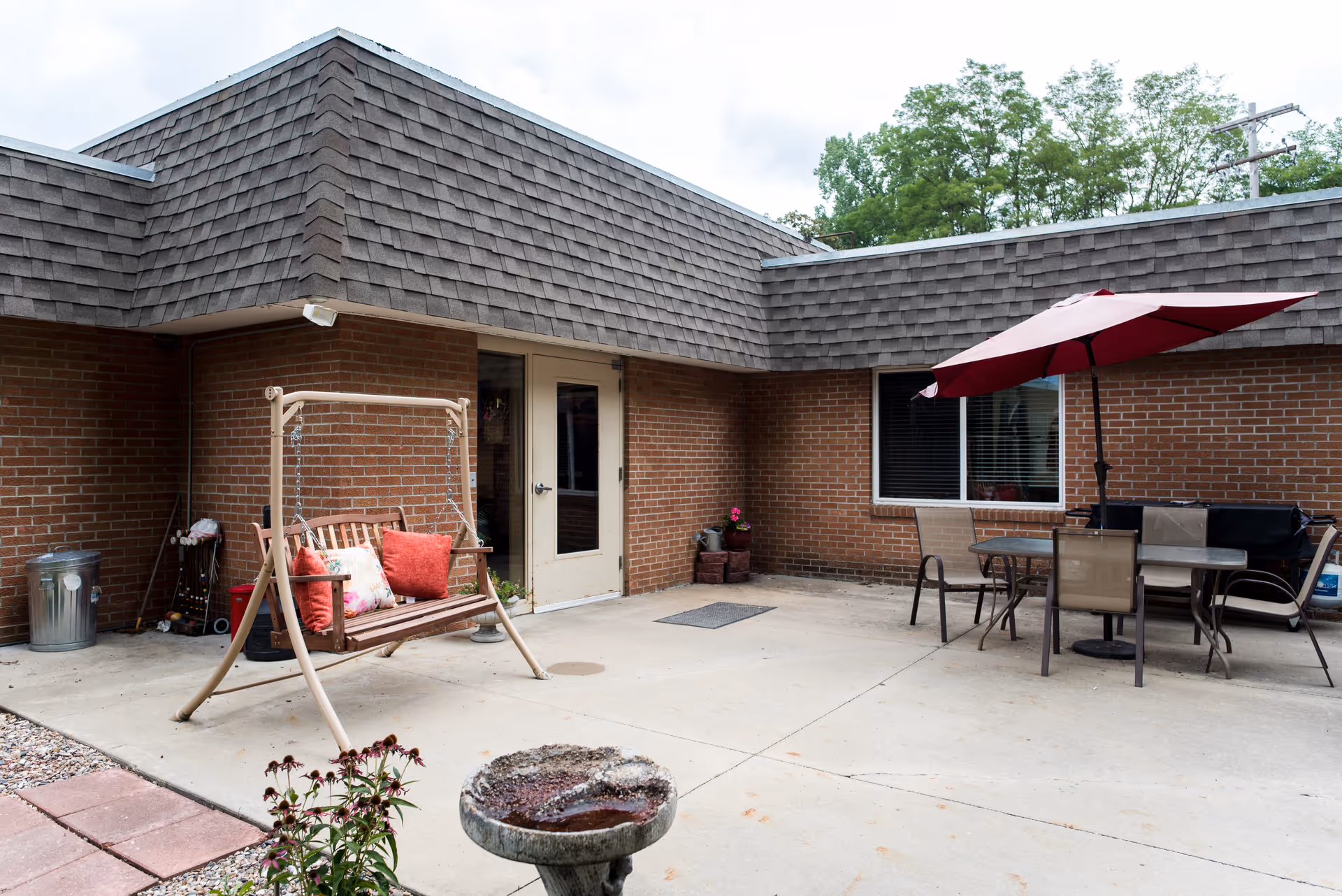 Outdoor patio area at Wathena Healthcare and Rehabilitation Center featuring a wooden swing with red and floral cushions, a table with four chairs under a red umbrella, a birdbath, and a brick building with a door and window in the background.