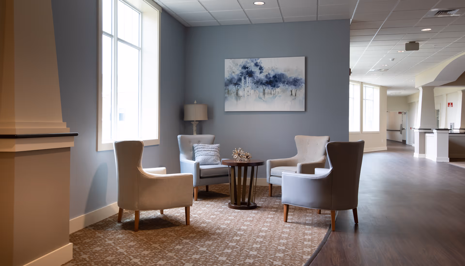 A cozy seating area in a senior living facility with four armchairs arranged around a small round wooden table. The walls are painted gray, and there is a large window letting in natural light. A modern painting with blue and white tones hangs on the wall, and a floor lamp is positioned in the corner. The floor has a patterned carpet in the seating area and wood flooring in the adjacent hallway.