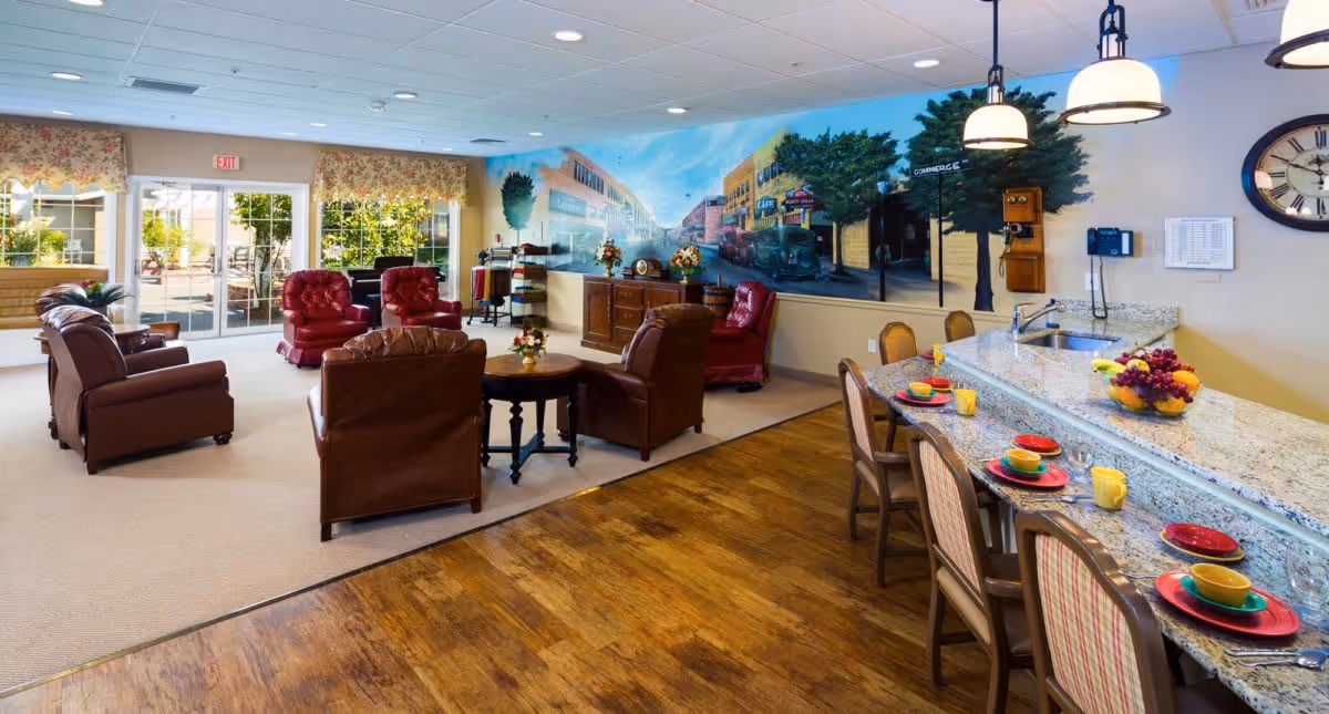 A spacious and well-lit common area in a senior living facility featuring comfortable leather armchairs arranged around small tables, a granite countertop with chairs set for dining, colorful dishware, and a large mural depicting a vintage street scene on the wall. Large windows with floral valances allow natural light to fill the room.