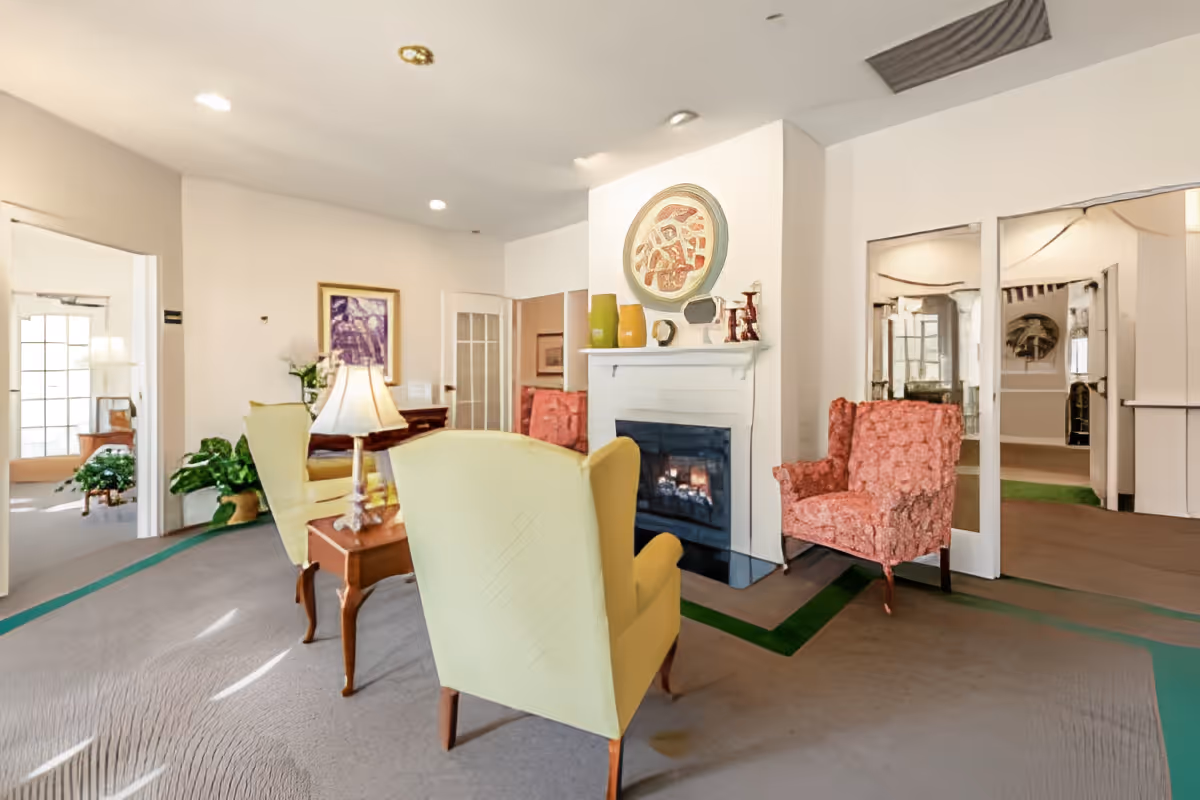 Bright communal sitting area with upholstered armchairs arranged around a lit fireplace and decorative wall art.