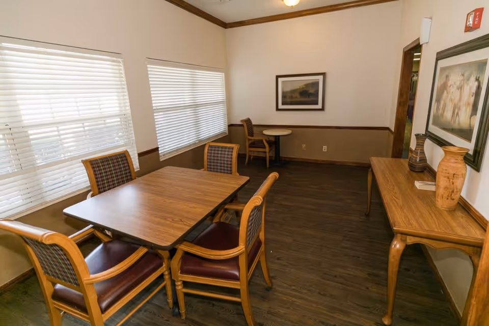 A small dining area with a wooden table and four chairs with plaid upholstery. There are two large windows with white blinds on the left wall. In the background, there is a smaller round table with two chairs. A wooden console table with decorative vases and framed artwork hangs on the wall to the right. The room has wood flooring and beige walls with a darker trim.