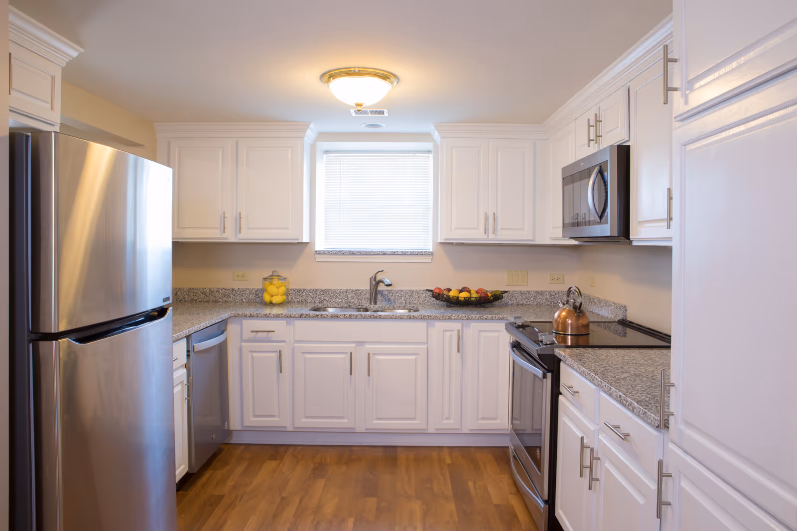 A bright kitchen with white cabinets, granite countertops, a stainless steel refrigerator, dishwasher, stove with a copper kettle, and a microwave. There is a window with blinds above the sink, and a bowl of fruit and a jar of lemons on the counter. The floor is wooden.