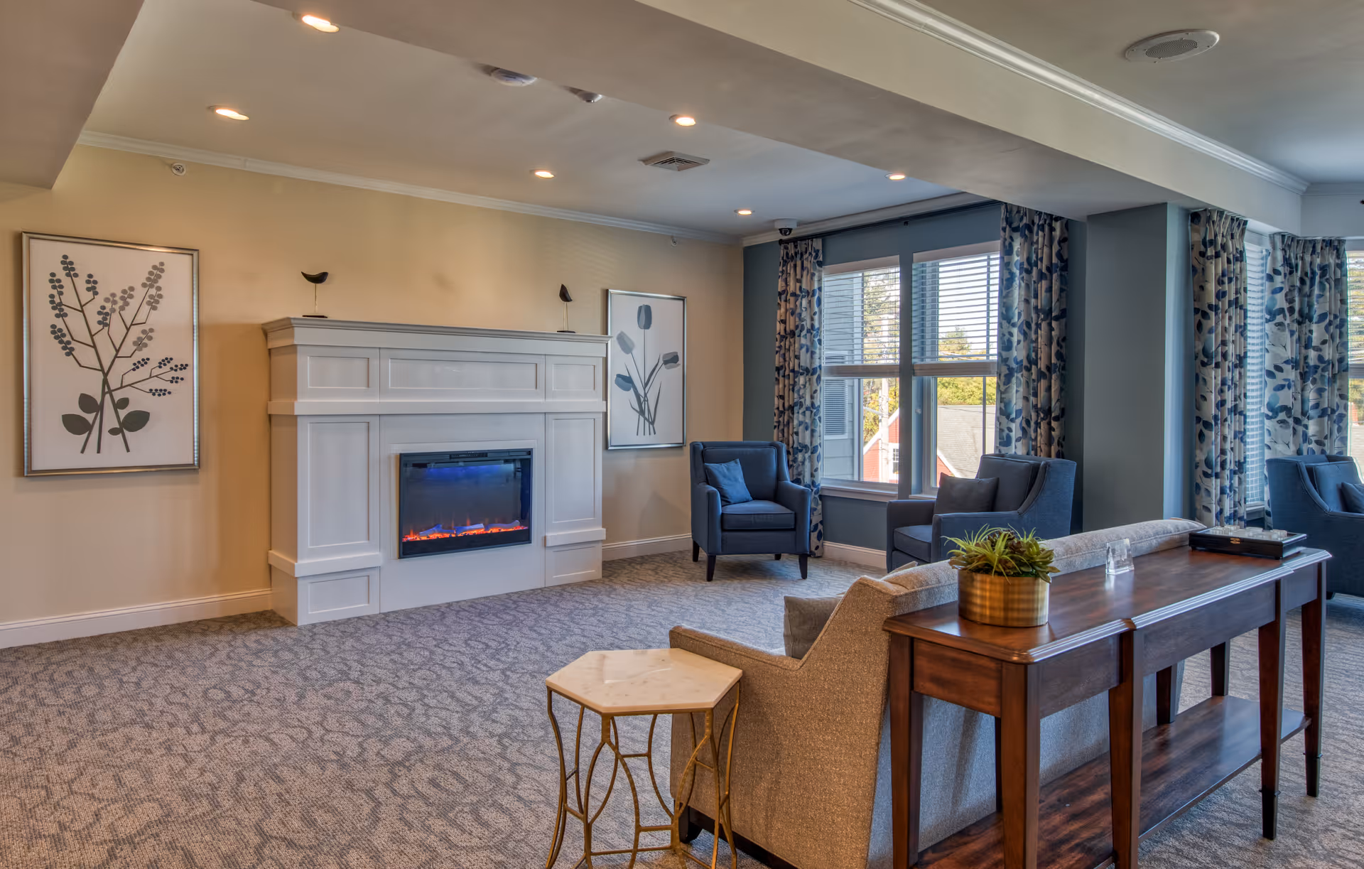 A cozy living room area in the Samuel F. Vilas Home featuring a white electric fireplace with blue and orange flames, two framed botanical prints on the wall, two blue armchairs near large windows with floral curtains, a beige sofa, a wooden console table with a small plant, and a small hexagonal side table.