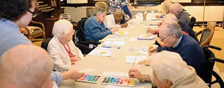 A group of elderly people sitting around a long table engaged in a painting activity, with paint palettes and brushes in front of them, in a well-lit room.