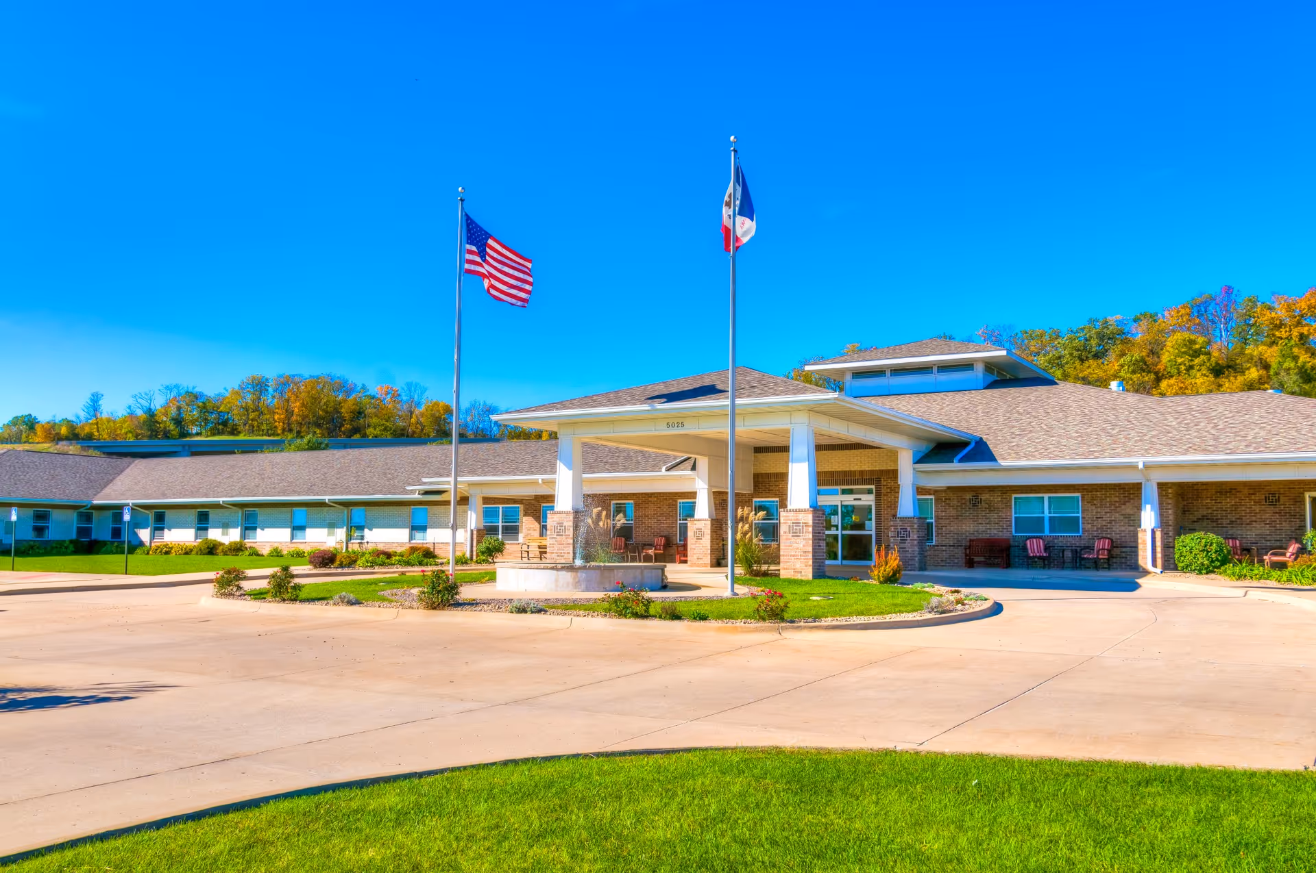 Exterior view of River Valley Place of Fort Madison, a single-story brick building with a covered entrance supported by white pillars. Two flagpoles stand in front, one with the American flag and the other with a state flag. The building is surrounded by a paved driveway, green lawn, and trees with autumn foliage in the background under a clear blue sky.