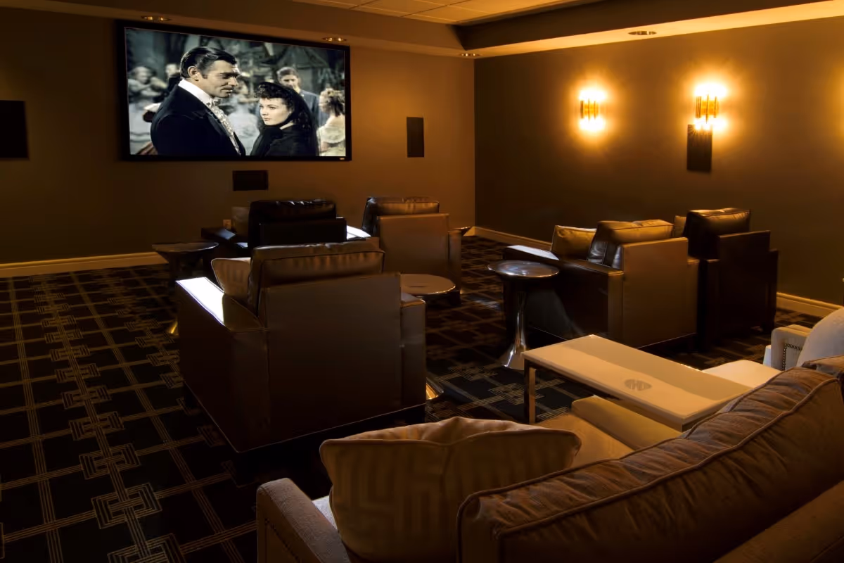 A cozy movie theater room with several leather armchairs and a sofa facing a large screen showing a black-and-white film. The room has patterned carpet, warm wall sconces, and a small table between the chairs.