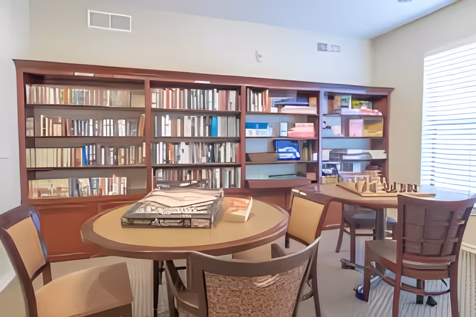 A cozy reading or activity room with round tables and chairs. One table has a chessboard set up, and another table has a tray with papers or magazines. A large wooden bookshelf filled with books and various items lines the back wall. A window with blinds lets in natural light.