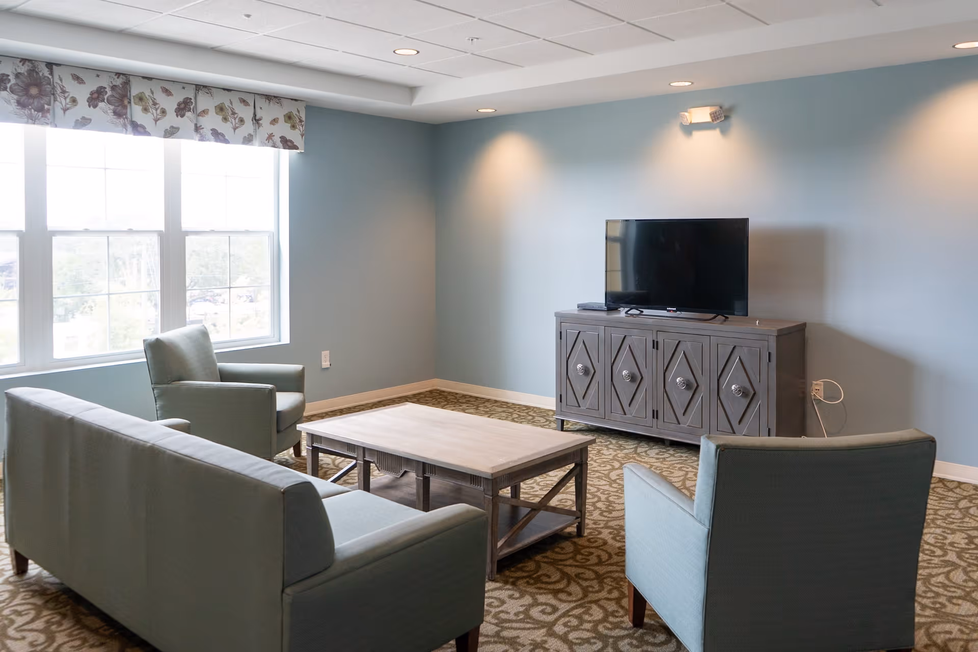A cozy living room with light blue walls and a patterned carpet. The room features a flat-screen TV on a decorative wooden cabinet, a light wood coffee table, a light blue sofa, and two matching armchairs. Large windows with floral valances allow natural light to fill the space.