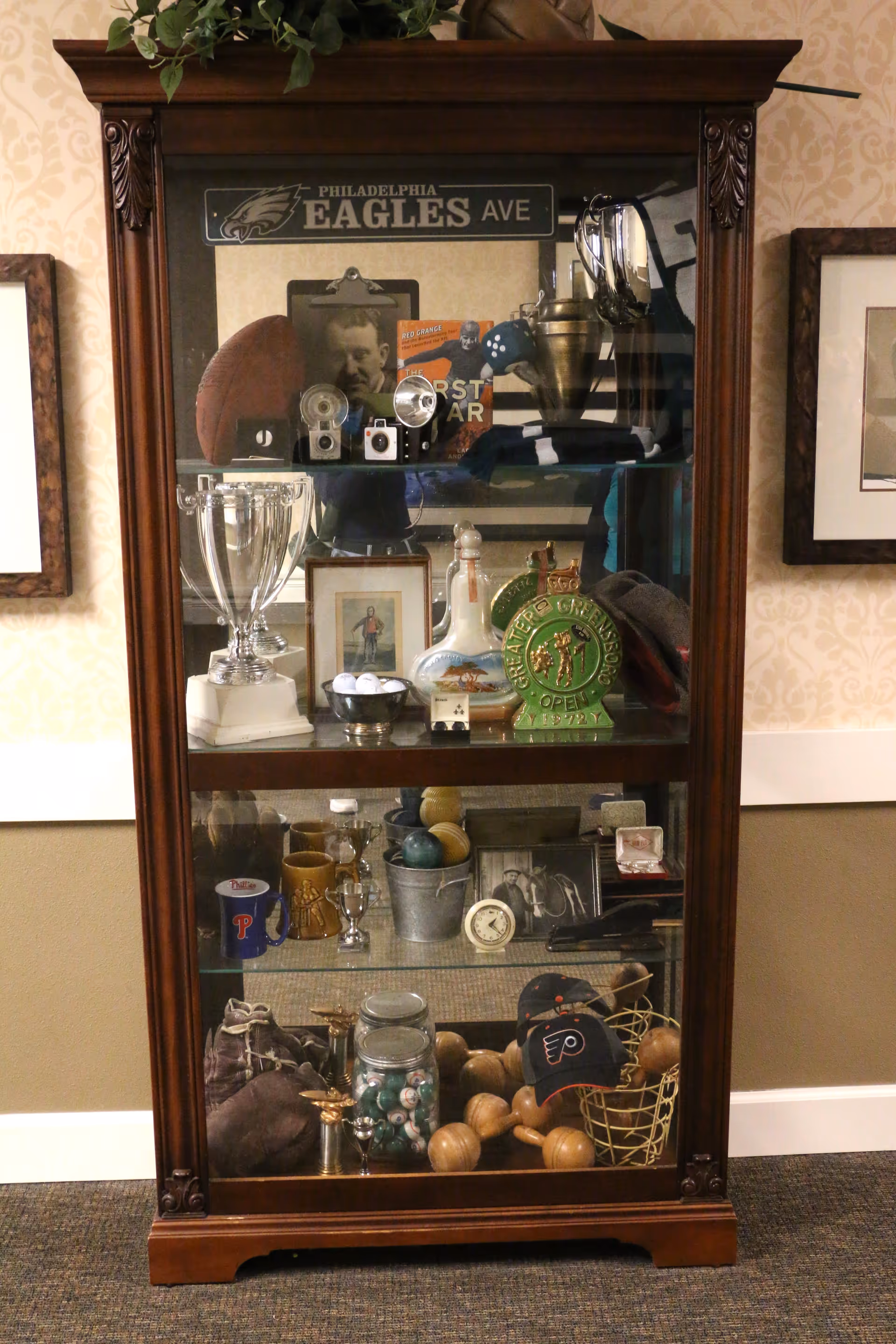 A wooden display cabinet with glass doors containing various vintage and sports memorabilia including trophies, old cameras, a football, golf balls, hats, mugs, framed photos, and decorative items. The top of the cabinet has a sign that reads 'Philadelphia Eagles Ave'.