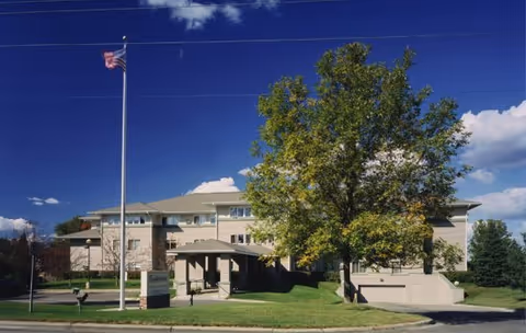 Exterior view of a multi-story senior living facility building with a large tree and an American flag on a flagpole in front. The sky is clear and blue with some clouds.