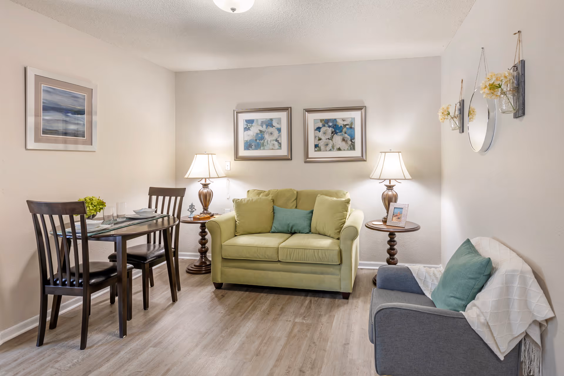 A cozy living room area with a light green loveseat adorned with three cushions, flanked by two wooden side tables each holding a lamp. To the left, there is a small dining table set with two chairs, plates, glasses, and a small plant centerpiece. On the right, a gray armchair with a green cushion and a white throw blanket is positioned against the wall, which is decorated with a round mirror and two small flower arrangements in hanging jars. The walls are light-colored and decorated with framed artwork.