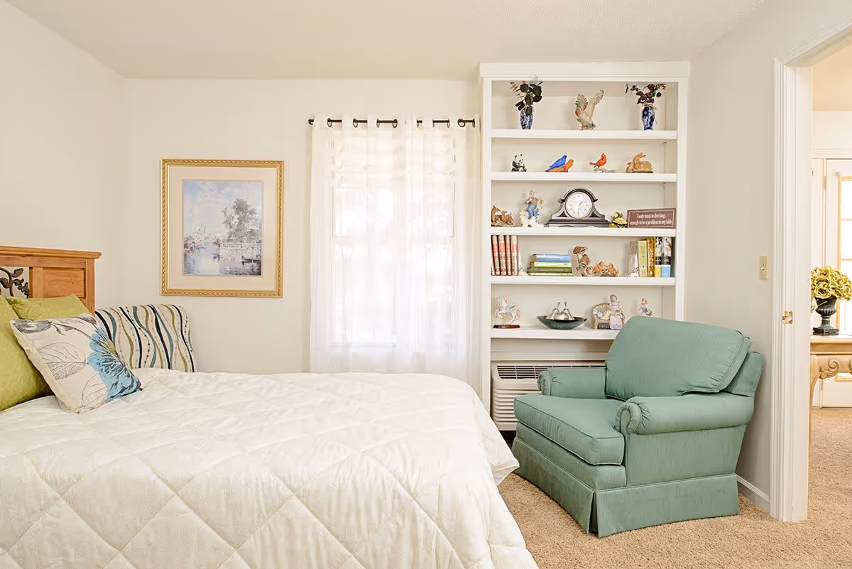 A cozy bedroom with a neatly made bed featuring white bedding and decorative pillows. Next to the bed is a green upholstered armchair. A built-in white bookshelf filled with various decorative items, books, and a clock is against the wall. A window with sheer white curtains lets in natural light, and a framed painting hangs on the wall beside the window. The room has beige carpeting and light-colored walls. An open doorway leads to another room with a table and a vase of flowers.