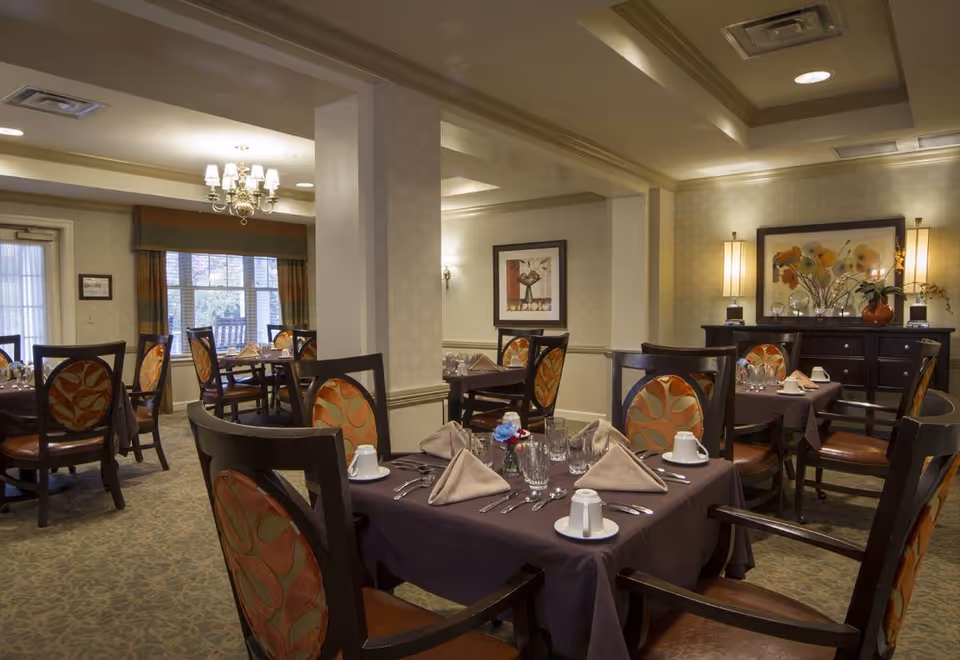 A warmly lit dining room in a senior living facility with multiple tables covered in dark tablecloths, each set with folded beige napkins, glassware, cups, and silverware. The chairs have wooden frames with patterned upholstery. The room features a chandelier, framed artwork, and decorative lamps on a sideboard against the wall.