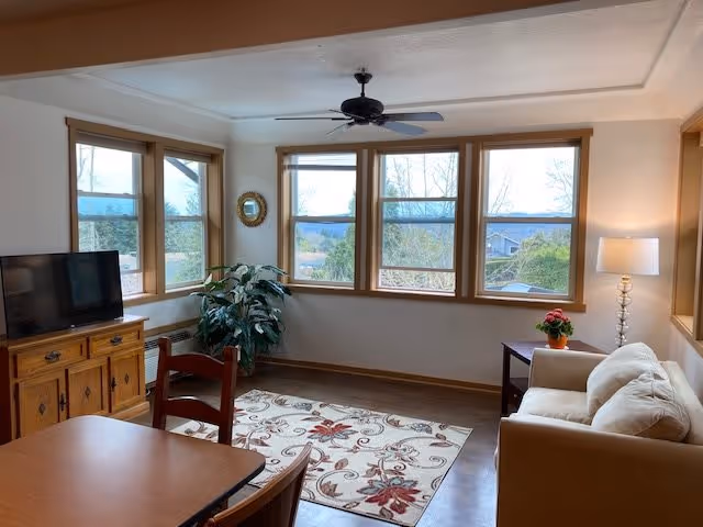 A cozy living room with large windows offering a scenic view of trees and hills. The room features a beige sofa with cushions, a wooden side table with a potted plant and a lamp, a wooden TV stand with a flat-screen TV, a wooden dining table with chairs, a decorative rug with floral patterns, a ceiling fan, and a large green plant in the corner.