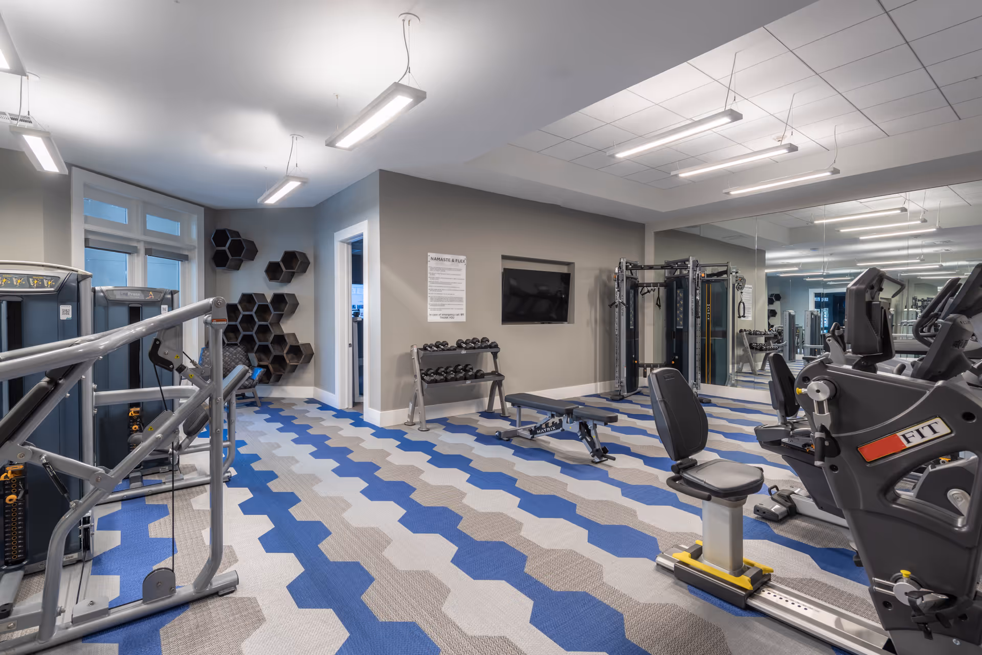 Bright, modern fitness room with exercise machines, weight bench, dumbbells, and blue-gray patterned flooring.