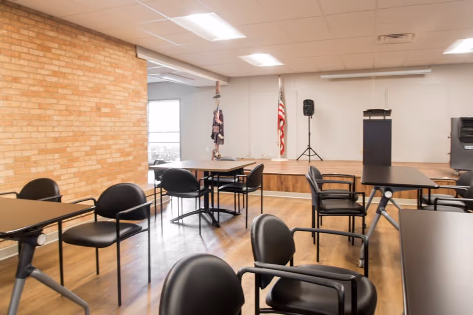 Interior room with multiple black chairs and tables arranged on a wooden floor. There is a small stage area with a podium, a speaker on a stand, and an American flag in the background. The walls are light-colored with a brick accent wall on the left side.