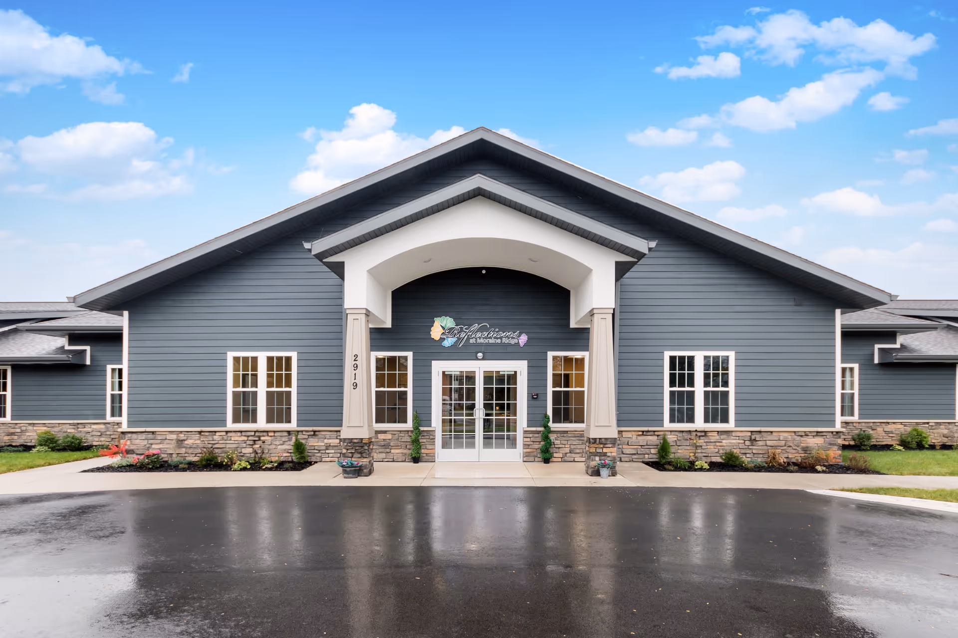 Front entrance of a single-story gray building with a covered portico, glass double doors, and landscaped beds.