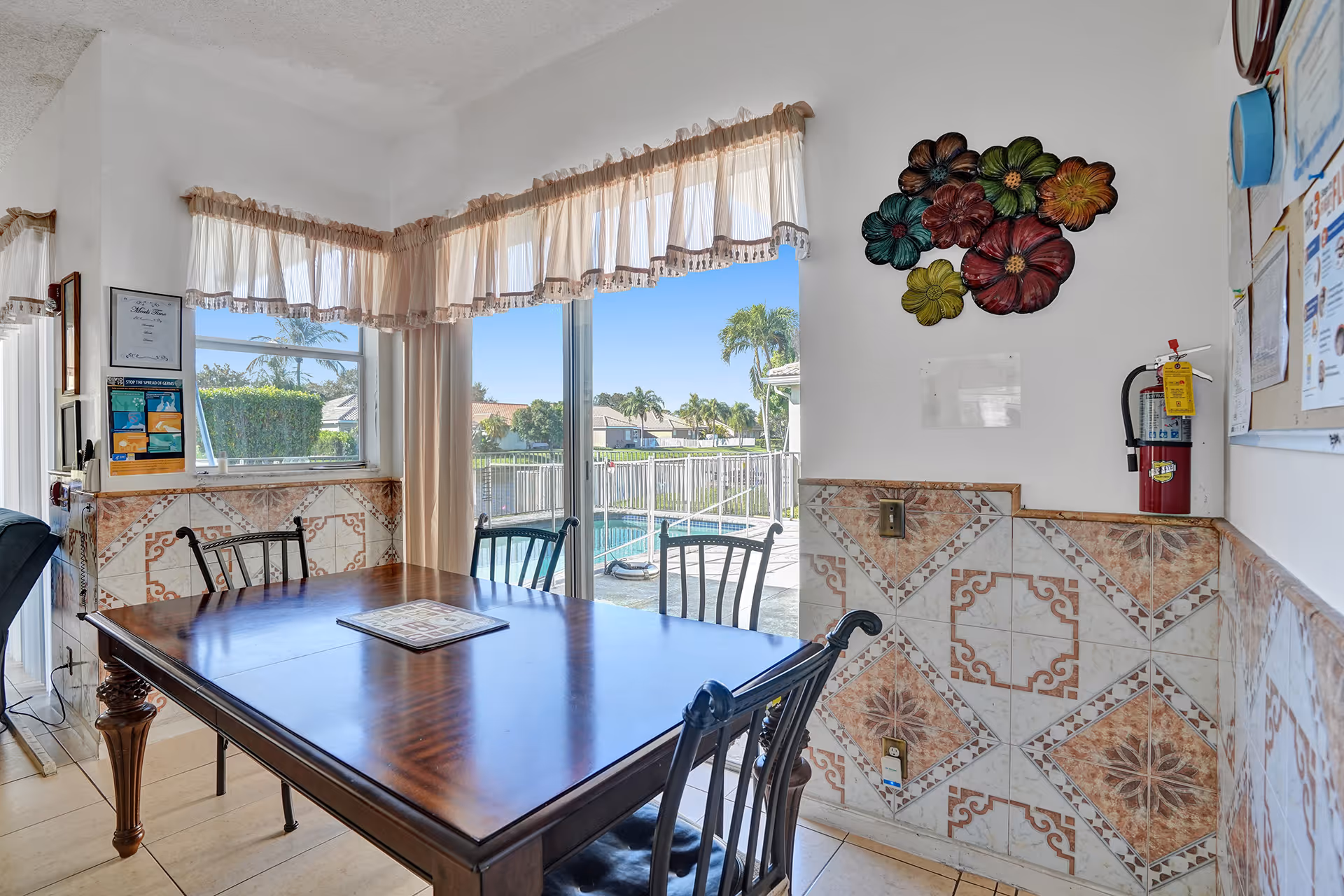A dining area with a wooden table and four metal chairs. The room has tiled walls with decorative patterns and a large window with sheer curtains that lets in natural light. Outside the window, a pool and palm trees are visible. There is a colorful flower wall decoration and a fire extinguisher mounted on the wall.