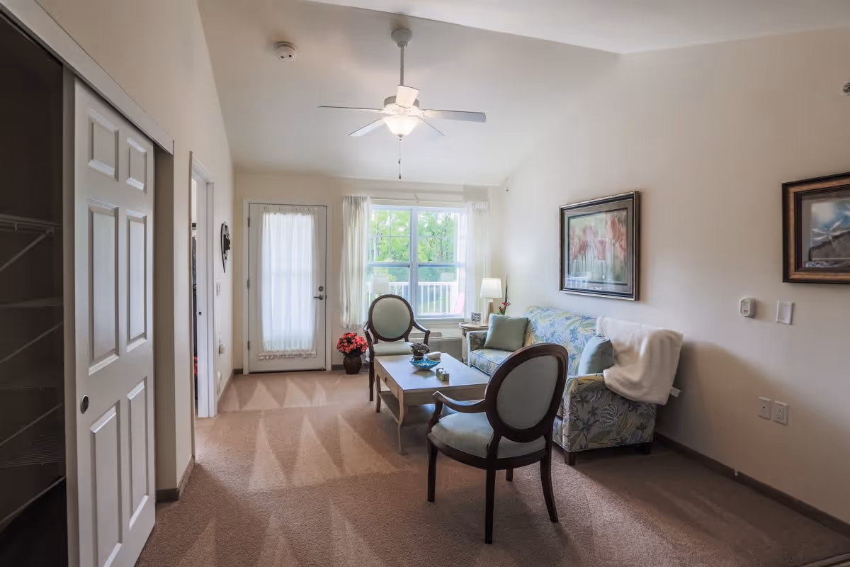 A cozy living room in a retirement facility featuring a floral-patterned sofa with a white throw blanket, two wooden chairs with light blue cushions, a wooden coffee table with decorative items, a side table with a lamp, framed artwork on the walls, a ceiling fan with light, a window with sheer curtains, and a door with a sheer curtain leading outside. The room has beige carpet and light-colored walls.