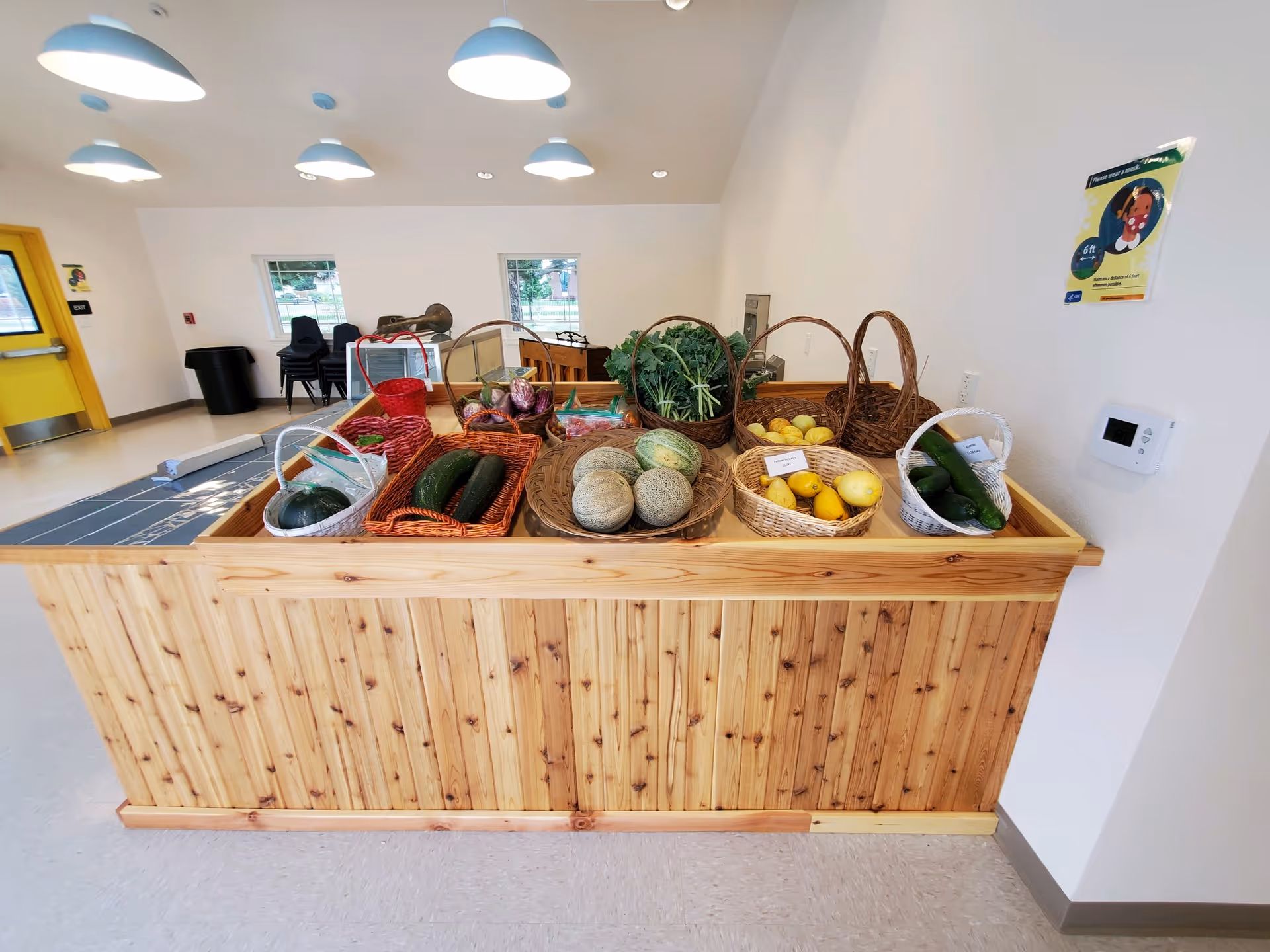 Interior view of a room with a wooden display counter holding baskets of fresh vegetables and fruits including cucumbers, cantaloupes, lemons, kale, and onions. The room has white walls, a yellow door, several blue pendant lights hanging from the ceiling, and windows in the background.