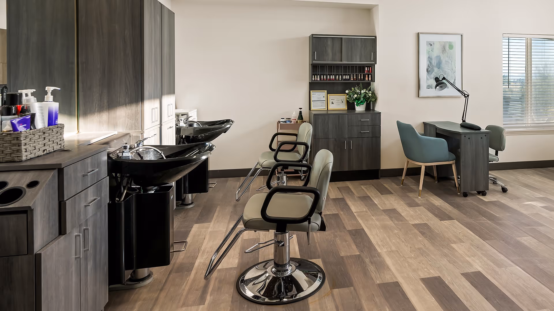 Interior view of a salon area in a senior living facility with two black hair washing sinks, two salon chairs, a cabinet with nail polish bottles, a small desk with a lamp, and two chairs near a window with blinds.