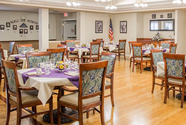 A dining room with multiple tables covered in white tablecloths and purple runners, each set with glassware, cups, and floral centerpieces. Wooden chairs with patterned cushions surround the tables. The room has light-colored wooden flooring, beige walls, and ceiling lights. An American flag is displayed in the corner, and a 'Wall of Honor' with framed photos is visible on one wall.
