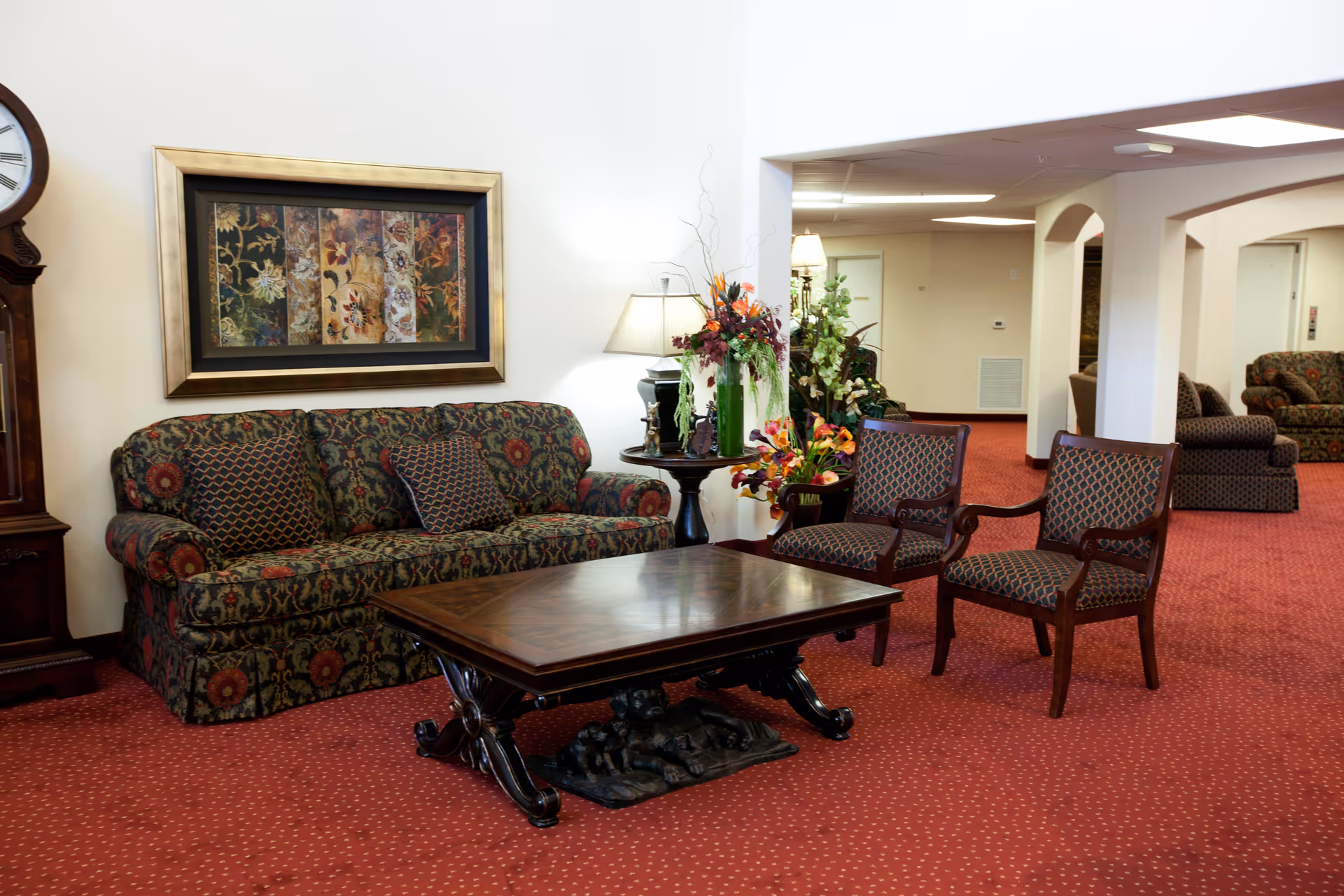 A cozy living room area in a senior living facility featuring a patterned sofa with matching cushions, two upholstered armchairs, a wooden coffee table with decorative base, a side table with a lamp and flower arrangements, a large framed artwork on the wall, and a grandfather clock. The room has red carpet flooring and white walls with archways leading to other areas.