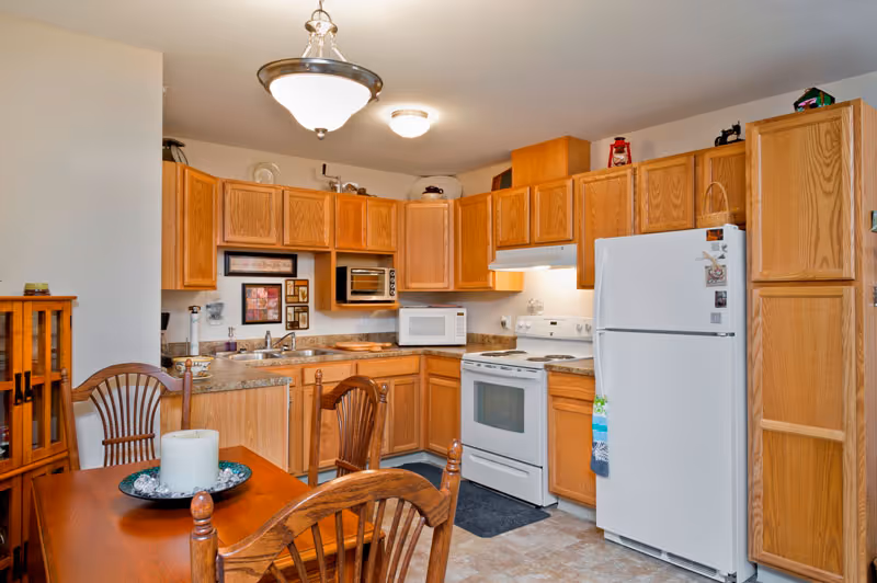 A kitchen with wooden cabinets, a white refrigerator, a white stove with an oven, a microwave, and a dining table with wooden chairs. The kitchen has overhead lighting and various kitchen items on the countertops.