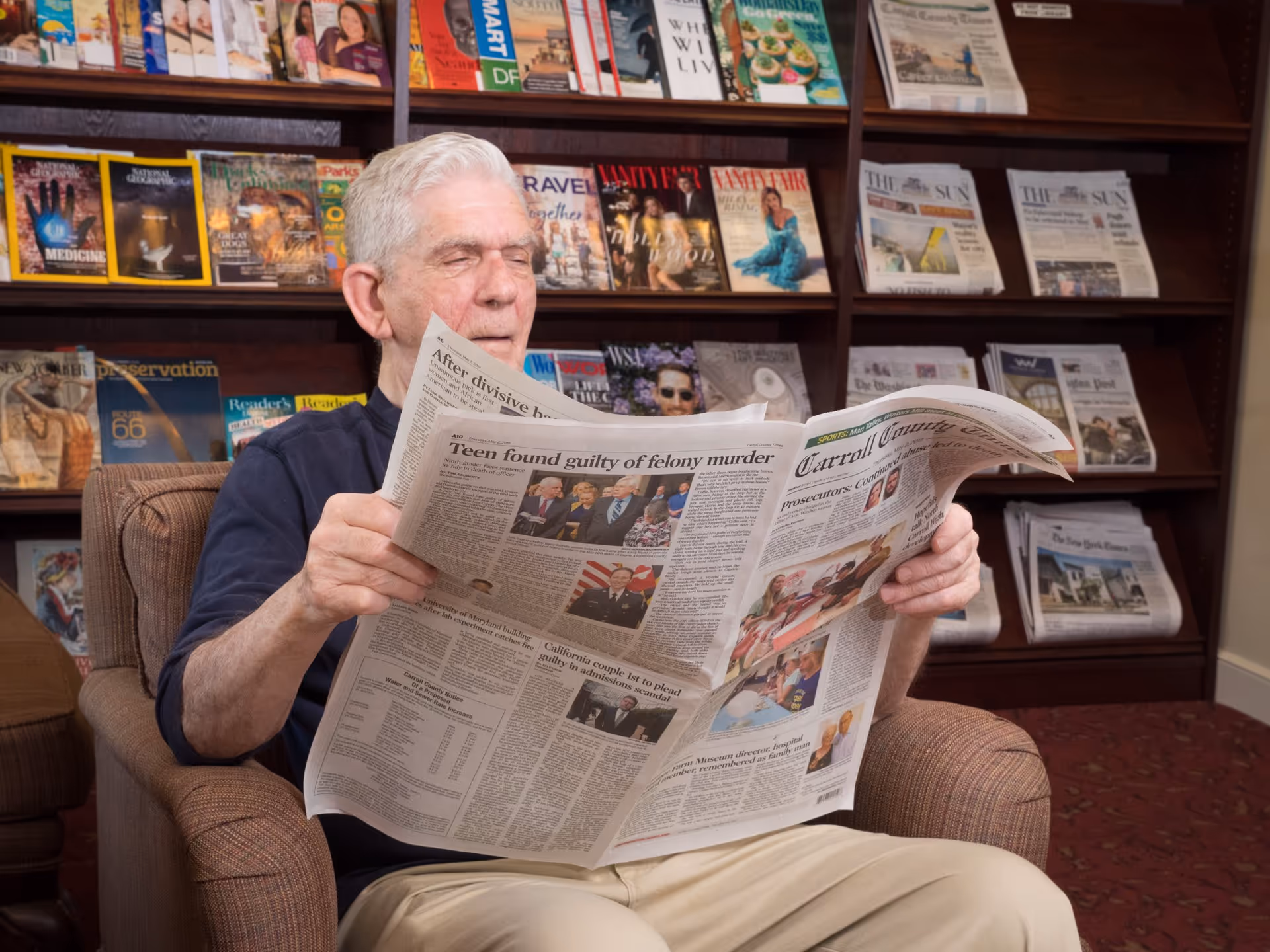 An elderly man with white hair sitting in a comfortable armchair reading a newspaper in a room with shelves filled with magazines and newspapers behind him.