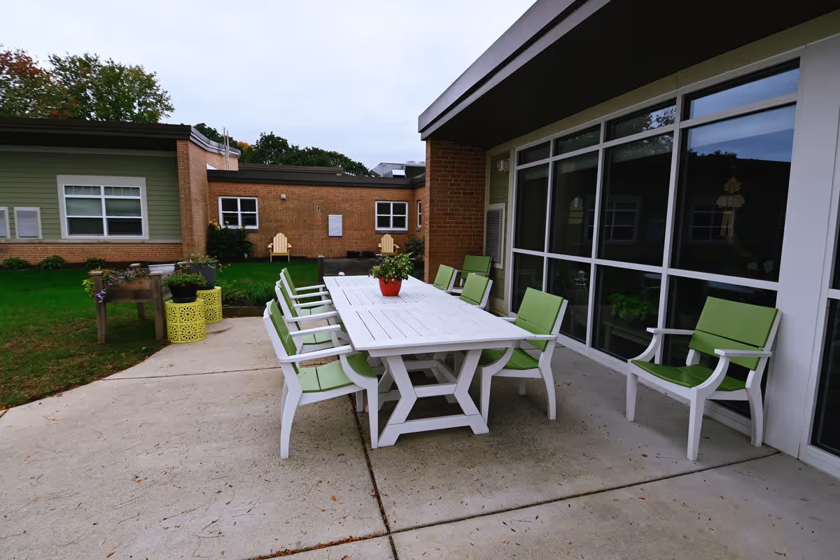 Outdoor patio area with a white rectangular table surrounded by eight white chairs with green cushions. The patio is adjacent to a building with large windows and brick and green siding. There are plants on the table and in the surrounding garden area, with two wooden Adirondack chairs visible in the background on a grassy lawn.