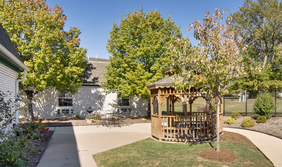 Outdoor patio area with a wooden gazebo surrounded by green trees and bushes. There is a concrete walkway and a small metal table with chairs near a white brick building under a clear blue sky.