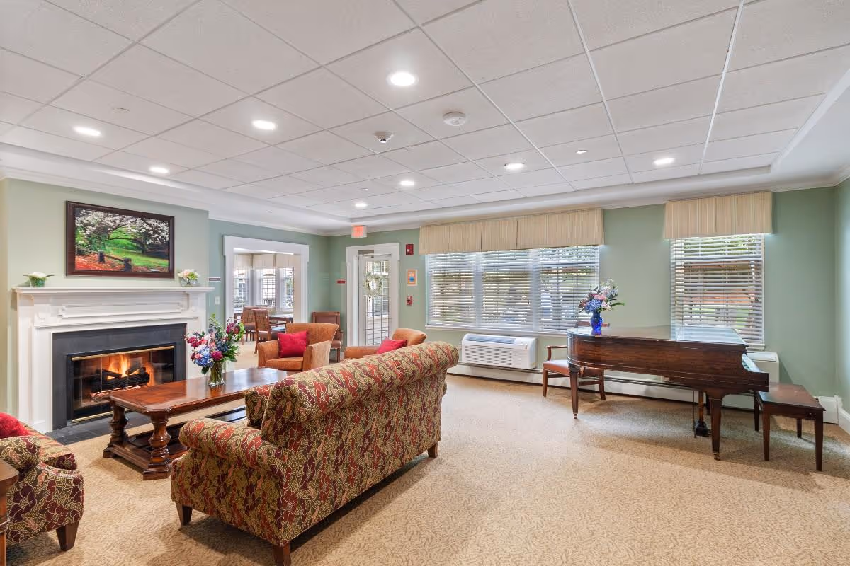 A cozy living room with a patterned sofa and armchairs arranged around a wooden coffee table in front of a lit fireplace. A grand piano with a vase of flowers on top is positioned near large windows with blinds. The walls are painted light green, and there is a framed picture above the fireplace. The room has a beige carpet and a white ceiling with recessed lighting.