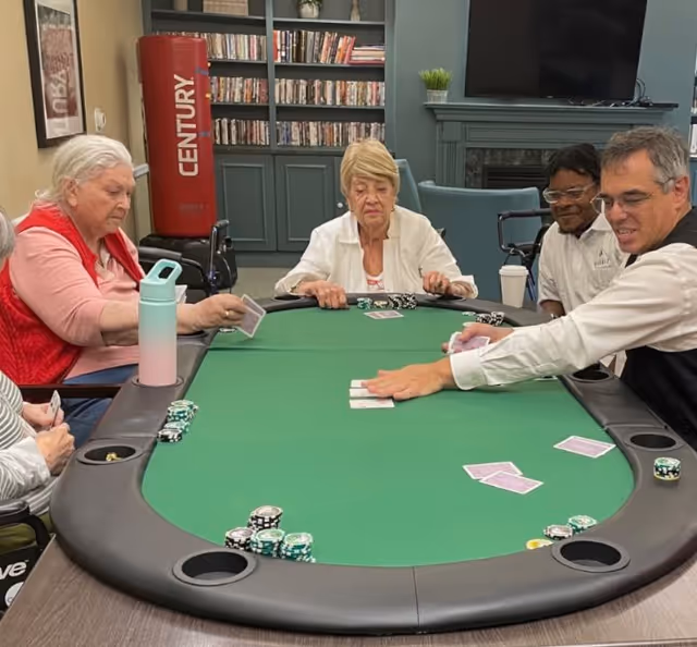 Four elderly individuals sitting around a green poker table playing cards and handling poker chips in a cozy room with bookshelves, a television above a fireplace, and a red punching bag in the background.