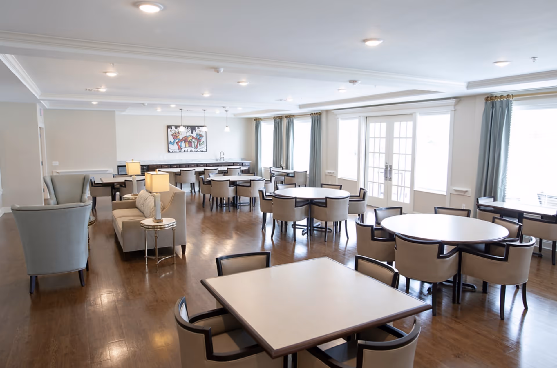Bright communal dining and lounge area with multiple round and square tables, upholstered chairs, and seating by large windows.