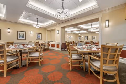 A spacious dining room with multiple round tables set with white napkins, plates, and silverware. The room features wooden chairs with cushioned seats, warm beige walls with framed artwork, and ceiling lights with decorative fixtures. The carpet has a red and brown floral pattern.