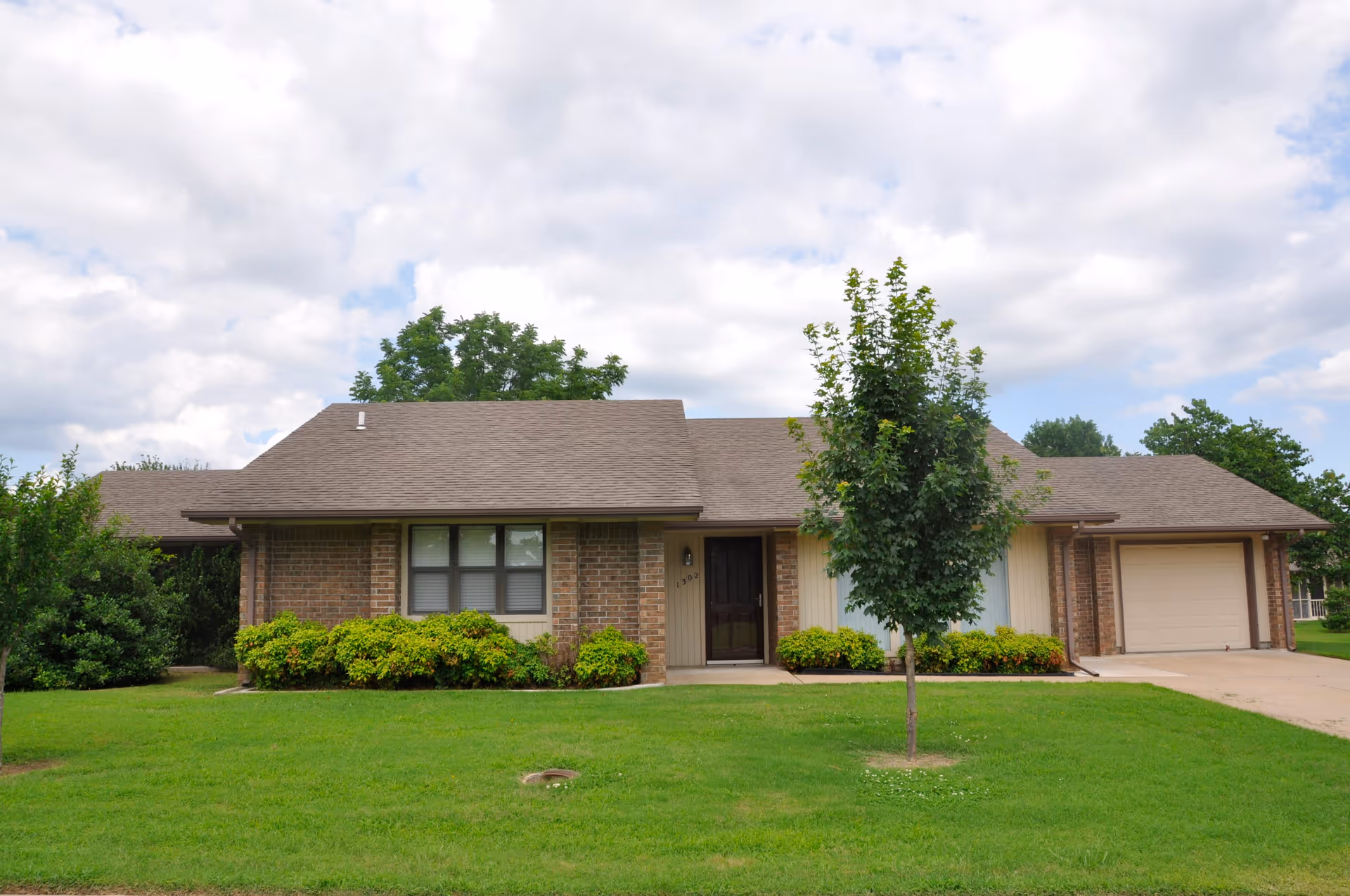 Single-story brick house with a brown shingled roof, a front door with the number 1302, a garage on the right side, green bushes along the front, a small tree in the lawn, and a partly cloudy sky overhead.