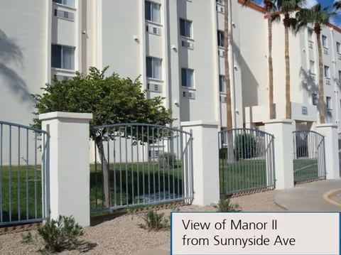 Exterior view of a multi-story white building with several windows, palm trees, and a metal fence with white pillars in front. There is a small landscaped area with grass and shrubs. A sign in the bottom right corner reads 'View of Manor II from Sunnyside Ave'.
