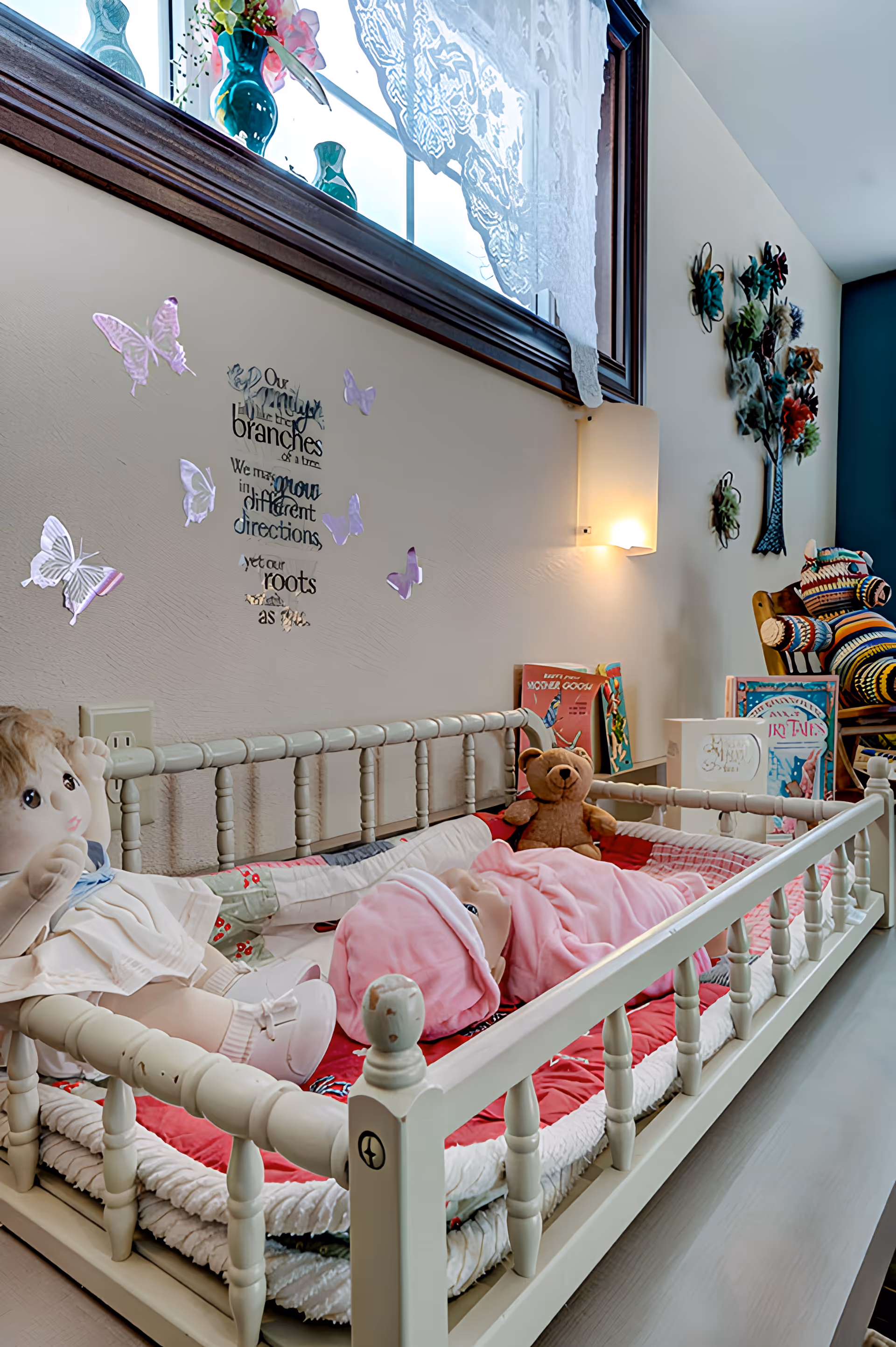 White daybed in a nursery with dolls, a teddy bear, pink bedding and butterfly wall decals beneath a window.