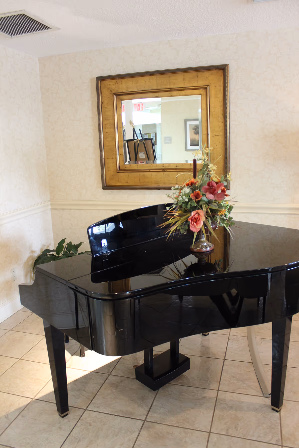 Black grand piano with a floral arrangement on top in a tiled interior room beneath a framed mirror.