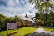 Brick community building with a pitched roof, front porch, walkway, lawn, trees, and a small directional sign.