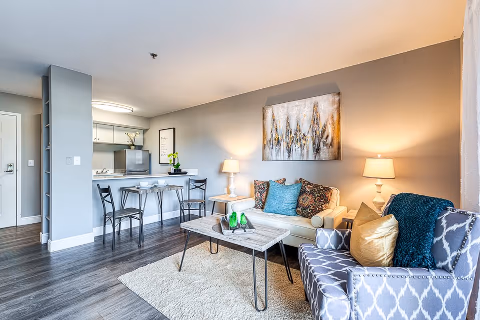 Bright furnished living room and kitchenette with a sofa, armchair, coffee table, dining counter and decorative artwork.