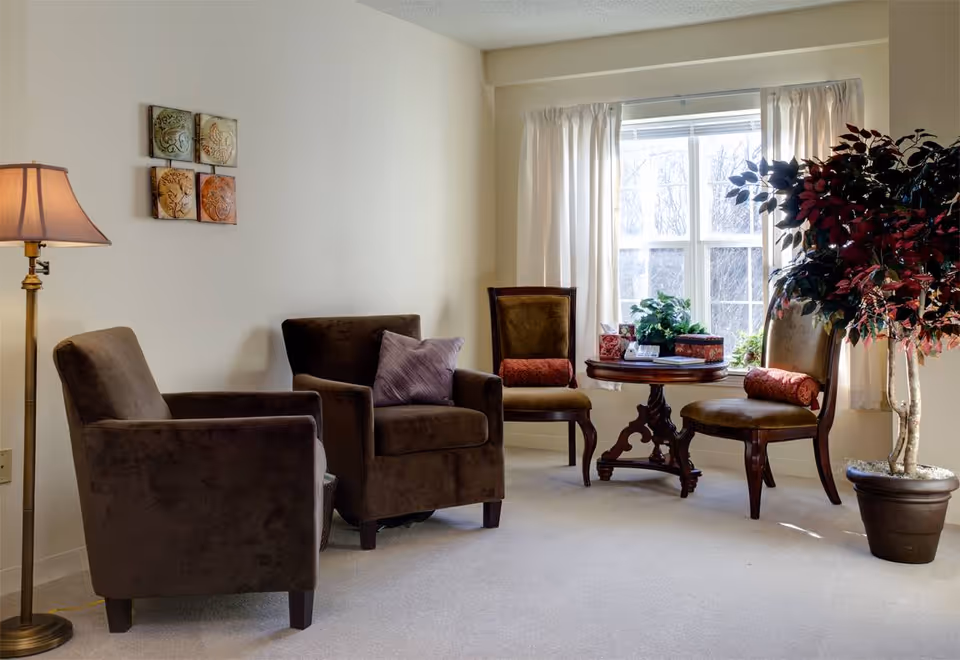 A cozy sitting area with two brown upholstered armchairs, two wooden chairs with red cushions, a round wooden table with plants and decorative items, a tall floor lamp, and a large potted plant near a window with white curtains.