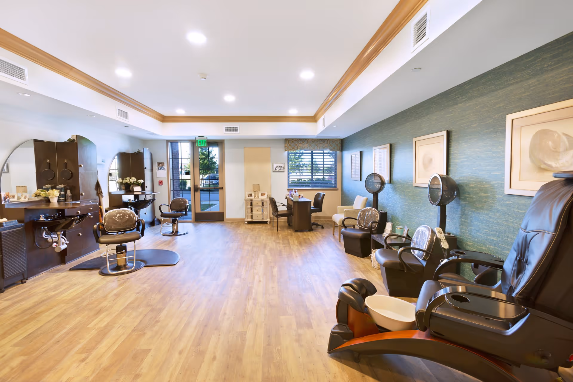 A spacious salon area with wooden flooring, featuring multiple styling chairs and mirrors on the left side, hair drying stations with chairs on the right, and a pedicure chair in the foreground. The room has a green accent wall with framed artwork, large windows, and a glass door letting in natural light.
