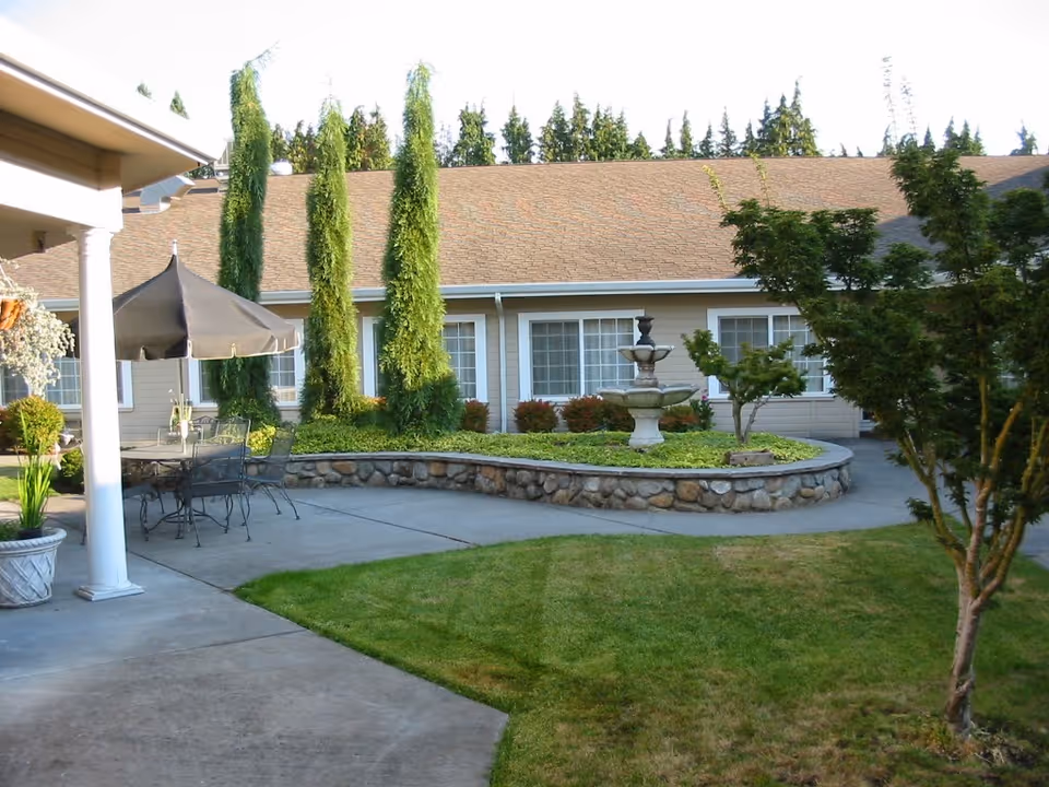 Outdoor courtyard area of a senior living facility with a stone-bordered circular garden featuring a tiered fountain, surrounded by greenery and trees. There is a patio with metal chairs and a table under a large umbrella, adjacent to a building with multiple windows and a sloped roof.