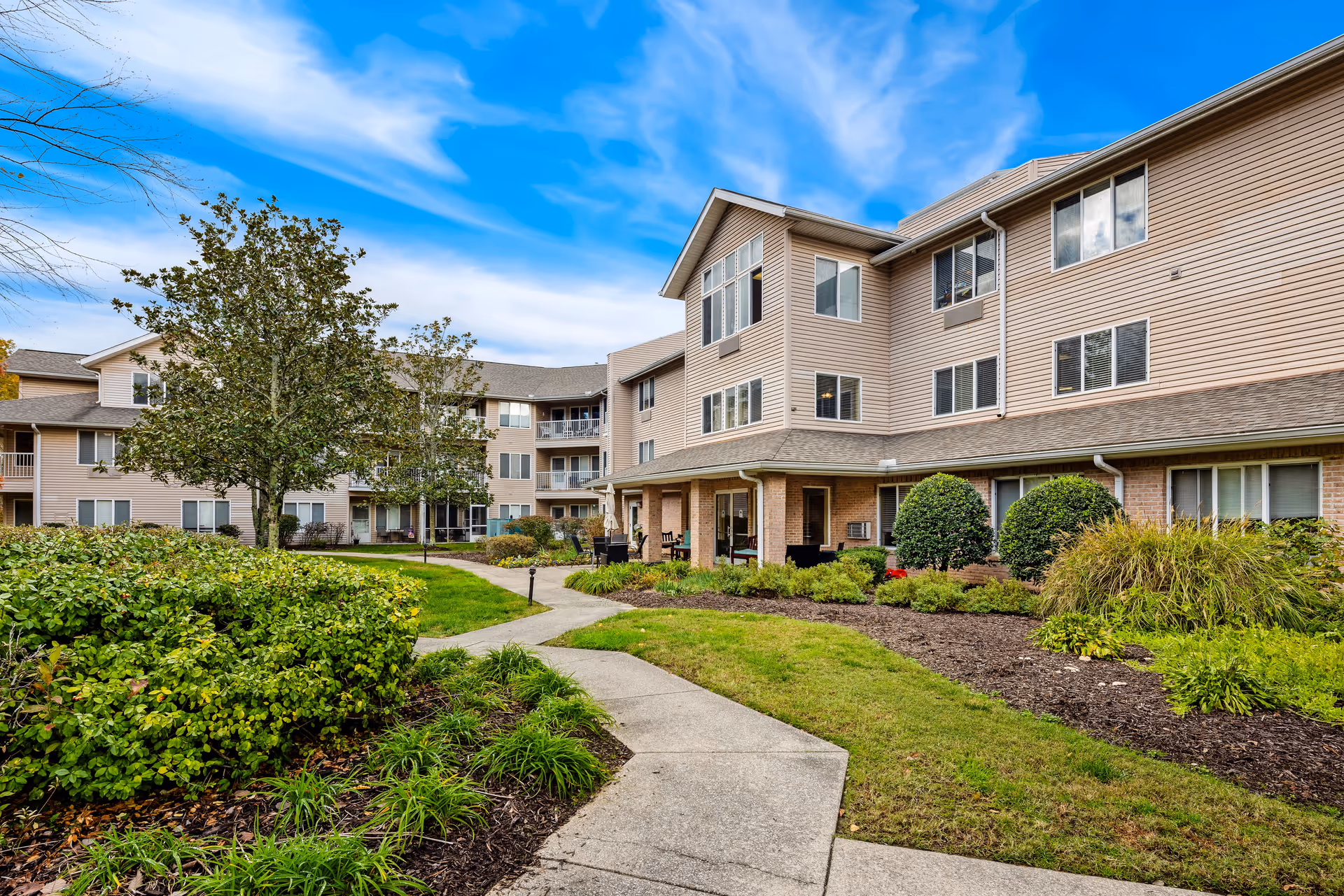 Exterior view of a three-story senior living community building with beige siding and multiple windows. A paved walkway curves through well-maintained green lawns and landscaped bushes and trees under a blue sky with some clouds.