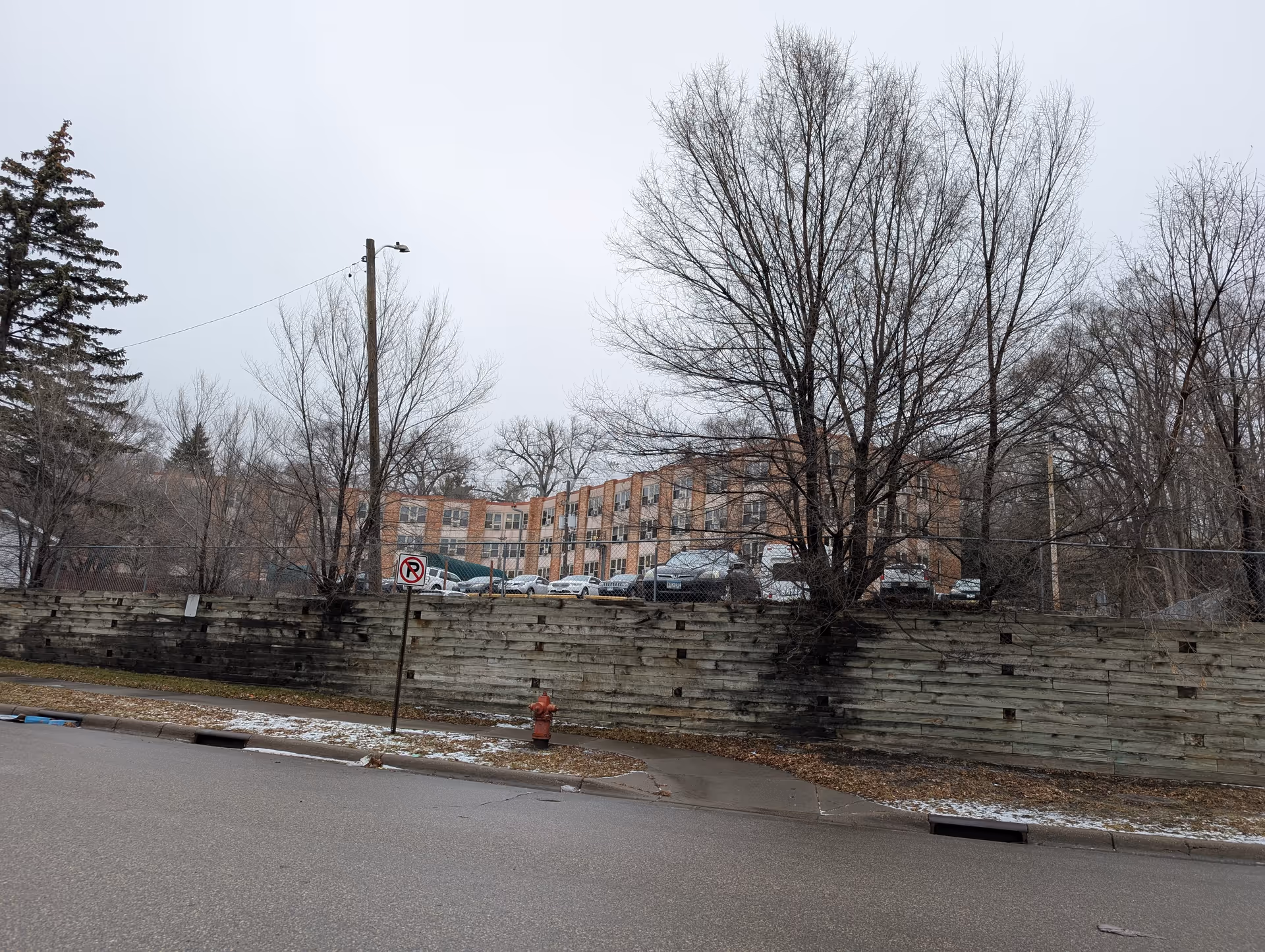 Exterior view of a multi-story brick building partially obscured by leafless trees and a wooden retaining wall, with a parking lot filled with cars behind a chain-link fence. A street with a no parking sign and a red fire hydrant is in the foreground under an overcast sky.