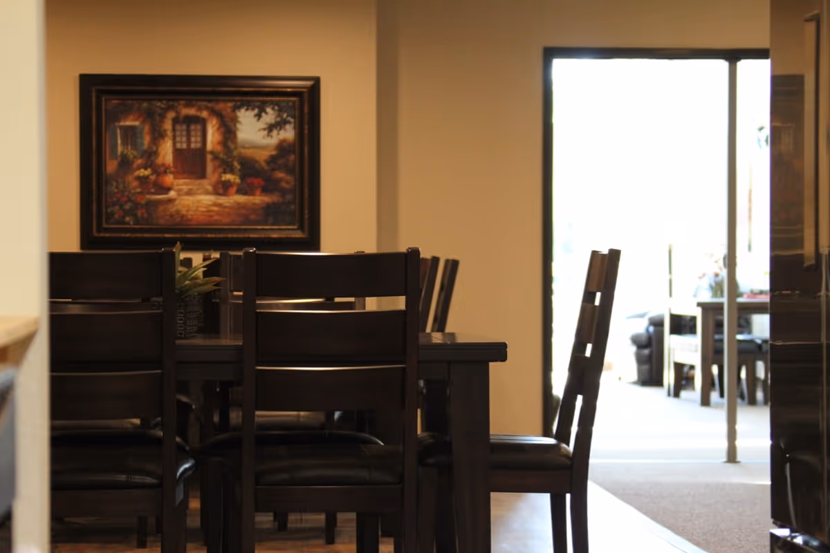 A dining area with a dark wooden table and chairs. A framed painting depicting a rustic doorway with potted plants hangs on the wall. In the background, there is a bright open doorway leading to another room with more furniture.